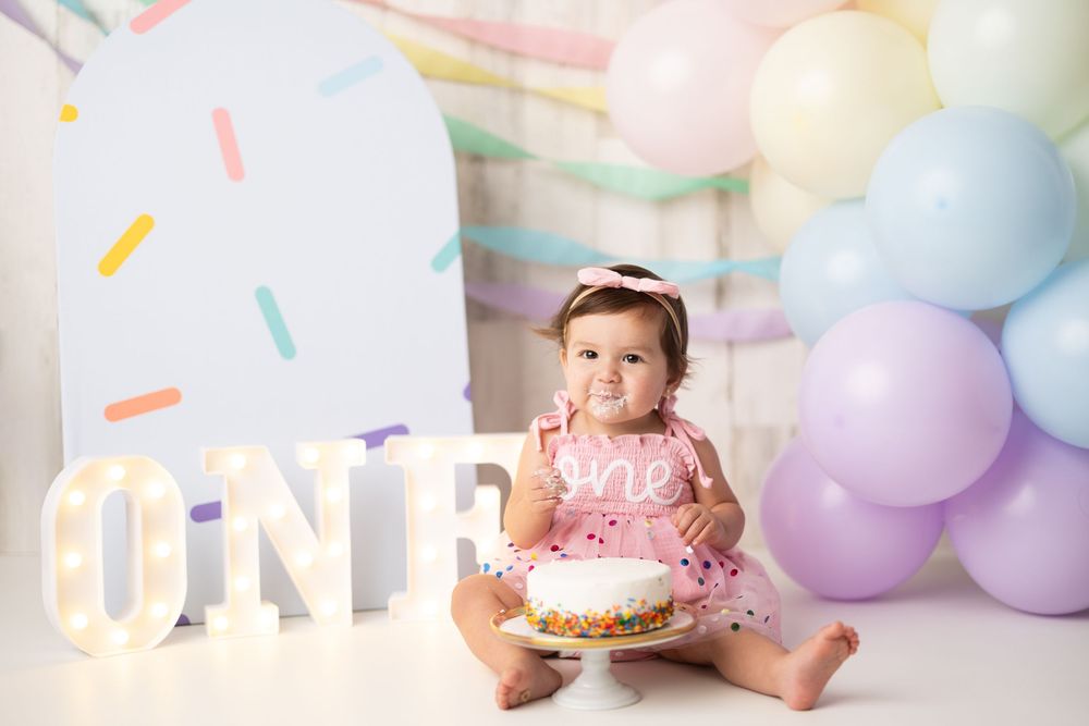 Baby girl in pink dress holds smash cake beside lit ONE sign and pastel balloons at first birthday photo shoot.