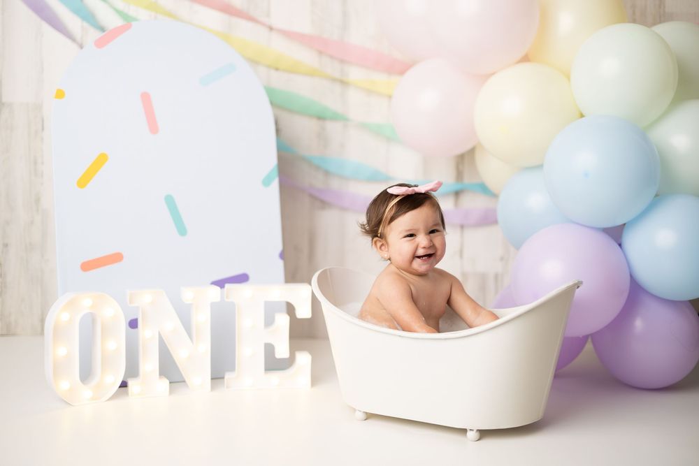 Baby girl laughing in white mini bathtub with ONE sign and pastel rainbow balloons at first birthday cake smash photo session.