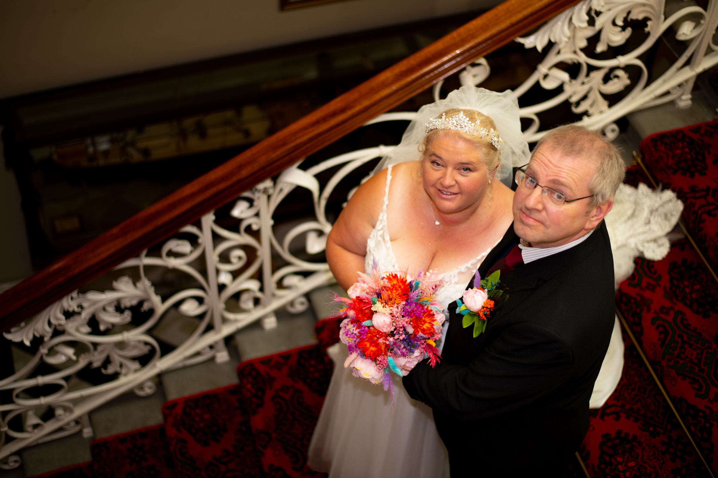 A wedding couple poses on elegant carpeted stairs with ornate white railings.