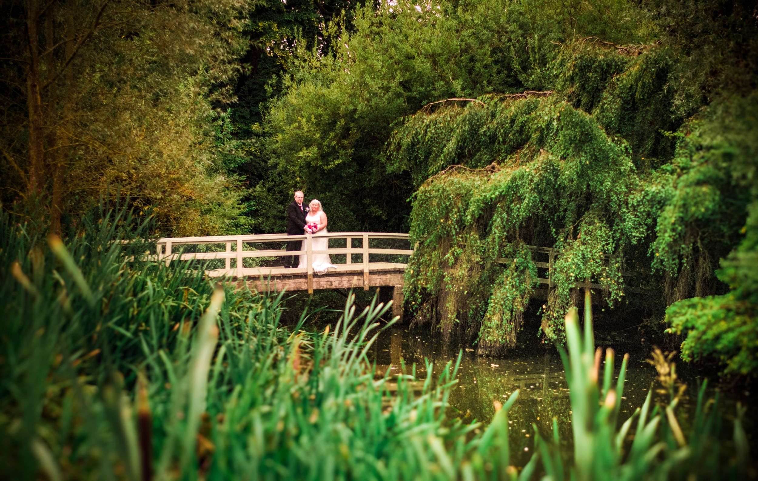 Romantic scene with a couple on a wooden bridge surrounded by lush gardens and weeping willows.