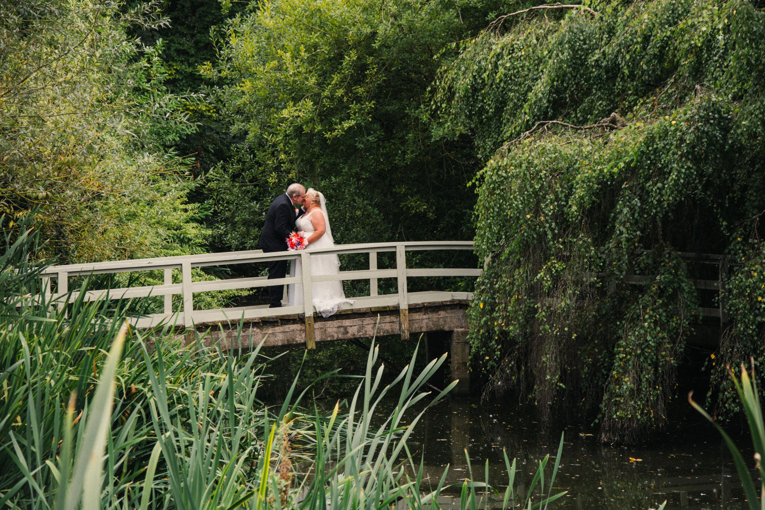 Couple shares romantic moment on wooden bridge surrounded by weeping willows and water plants.