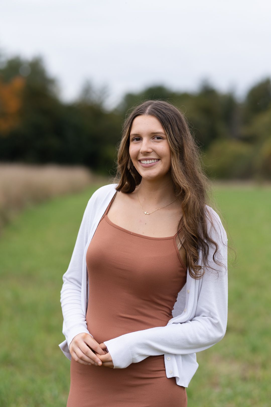 Woman in brown dress and white cardigan stands in natural field setting.