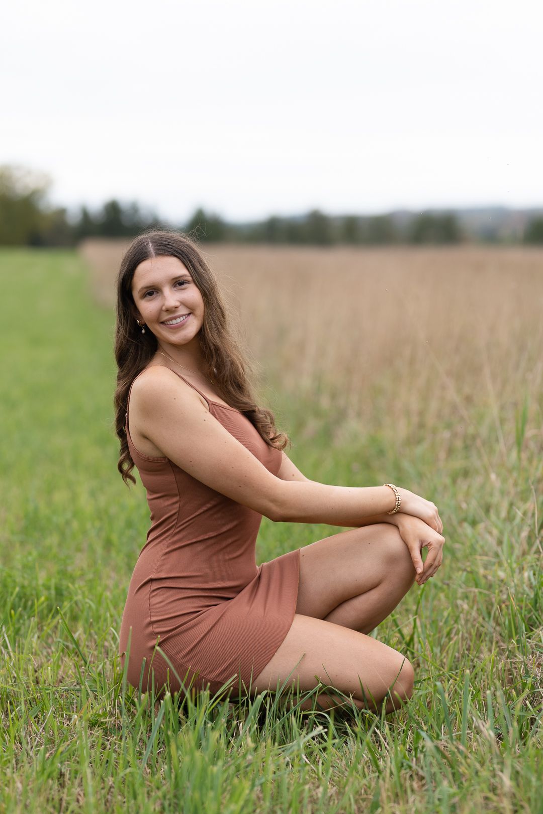 Woman in brown dress poses in tall grass field during outdoor photoshoot.