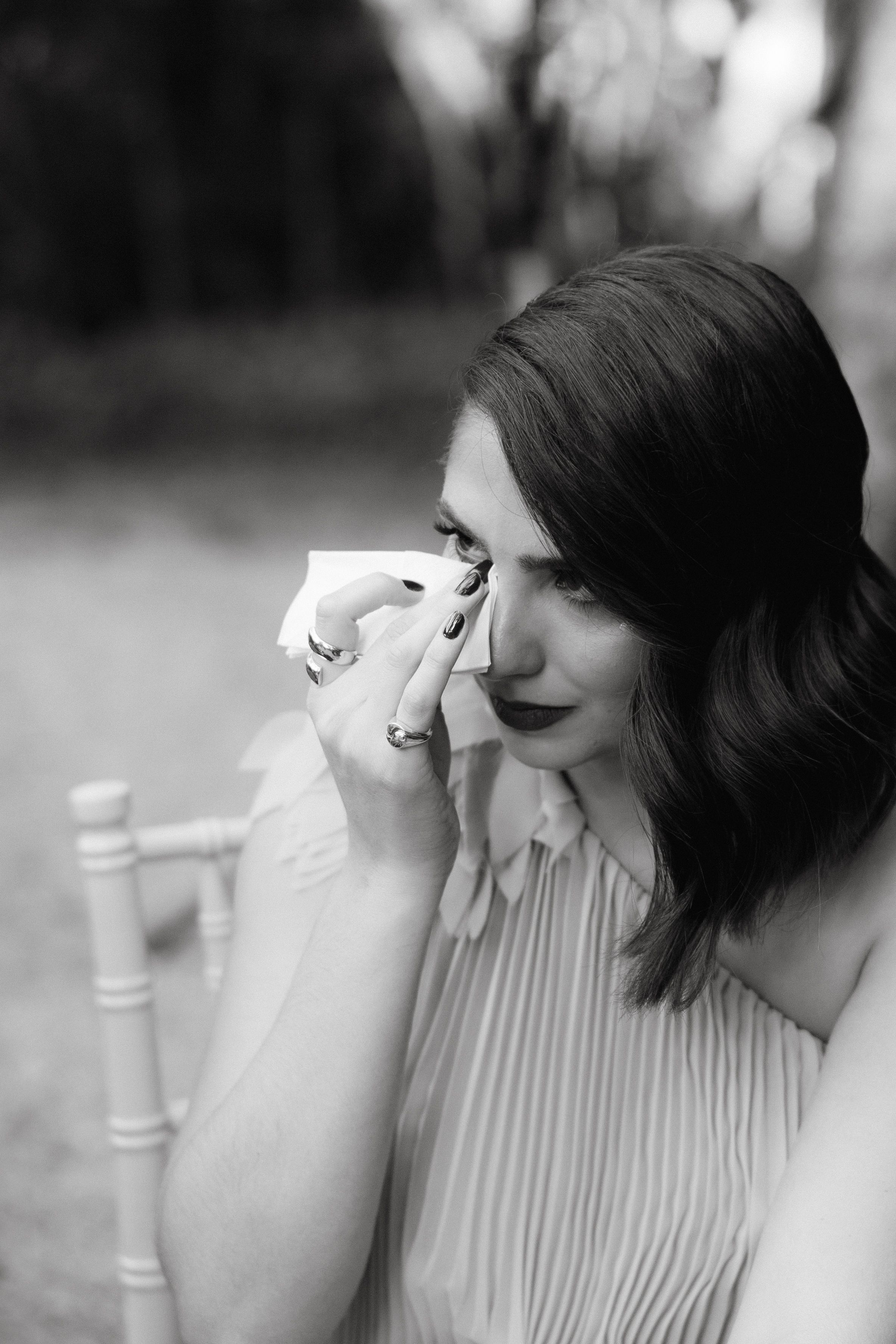 Black and white portrait of a woman in pleated dress wiping tears with a tissue during an emotional moment outdoors.