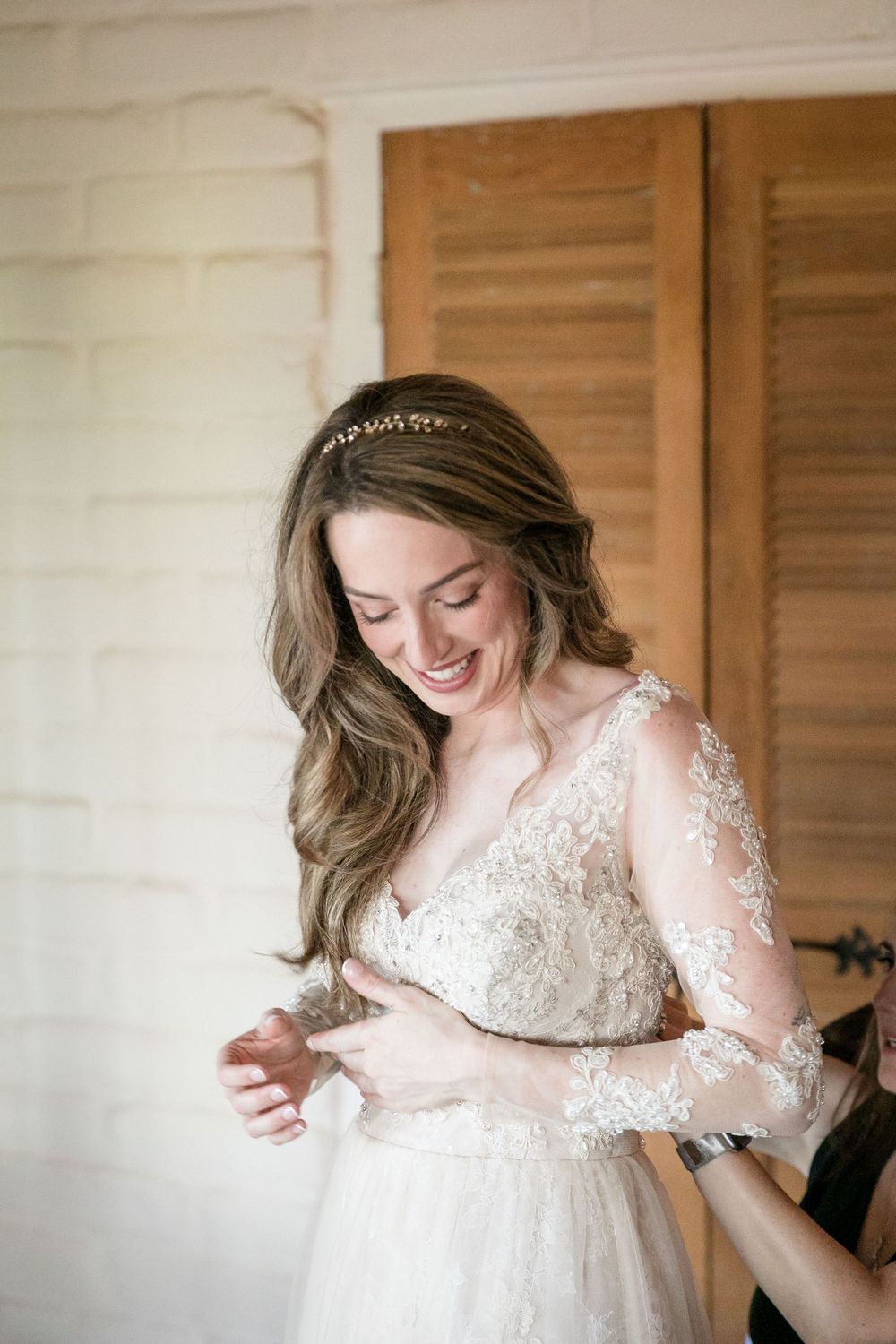 Smiling bride in lace wedding gown with sheer sleeves and floral headpiece getting dressed before her wedding ceremony.