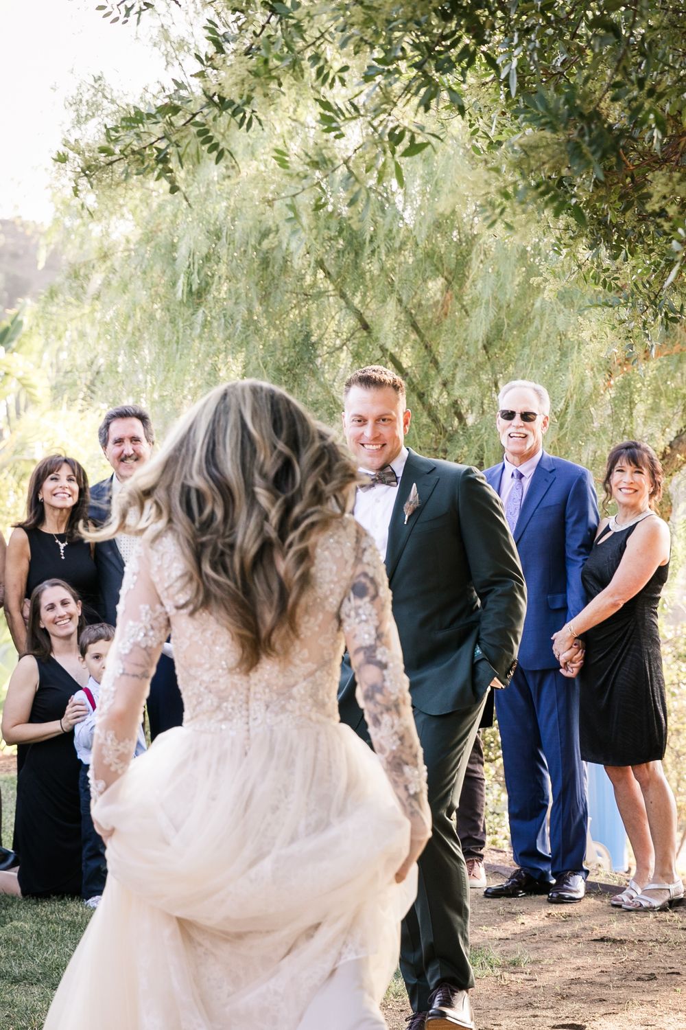 Bride in lace gown walks toward smiling groom in dark suit during outdoor wedding ceremony with guests watching.