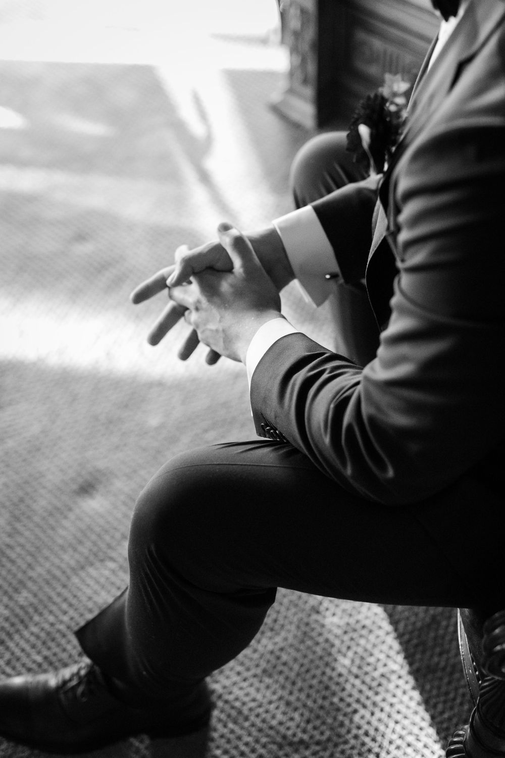 Black and white photo of a groom in a suit adjusting his cufflinks while seated, with dress shoes visible.