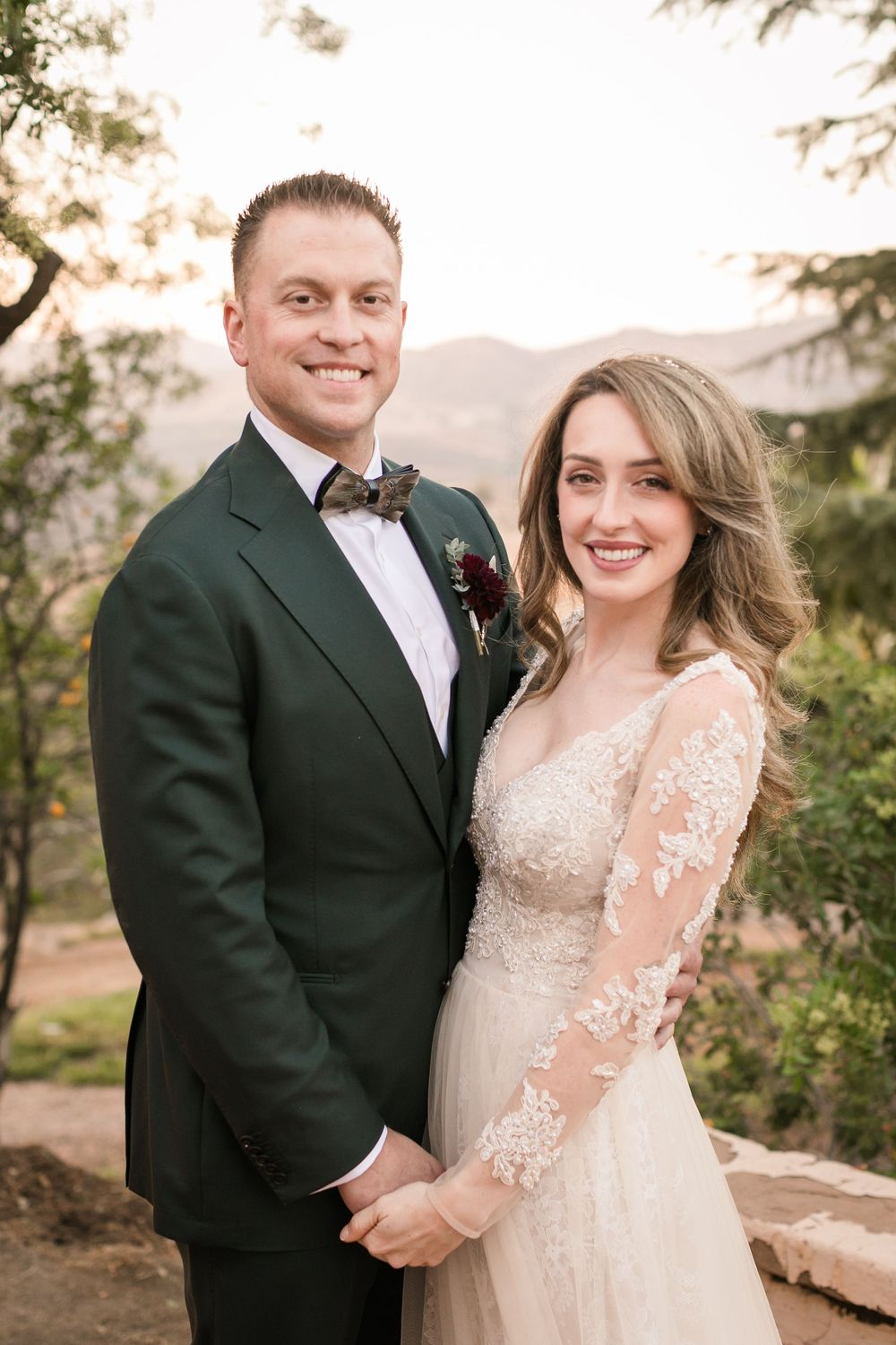 Smiling bride and groom pose outdoors in wedding attire with mountain backdrop at golden hour.