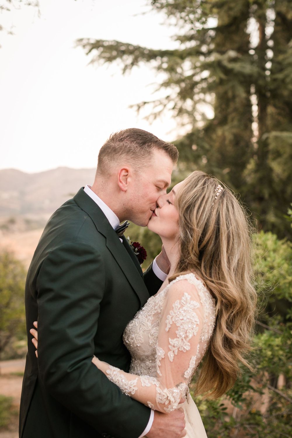 Bride and groom share a romantic kiss outdoors, surrounded by trees and rolling hills, bride in lace gown, groom in dark suit.