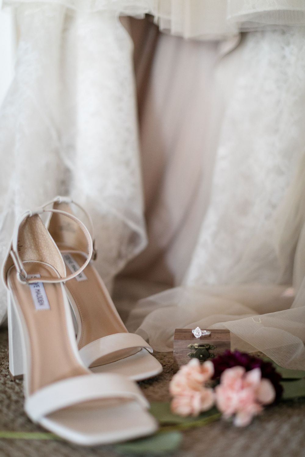 White strappy heeled wedding shoes beside pink flowers and a diamond ring box, with a lace bridal gown in the background.