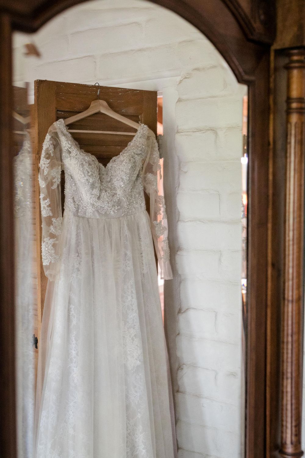 Elegant lace wedding dress with long sleeves hanging on a wooden door in a rustic white brick room.