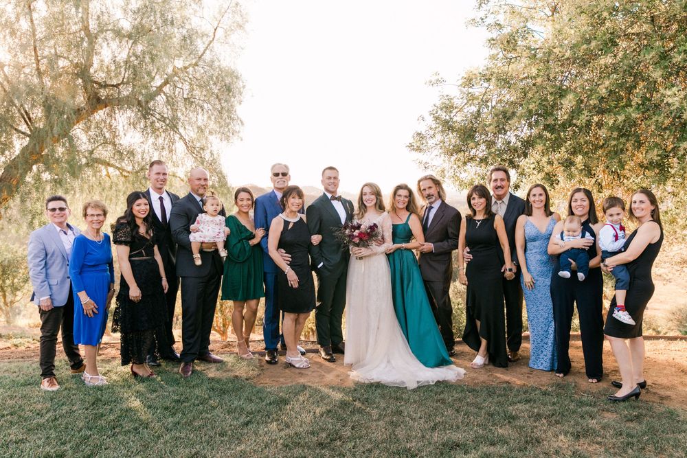 Wedding party group photo outdoors under trees, bride in white gown with teal sash surrounded by family in formal attire.