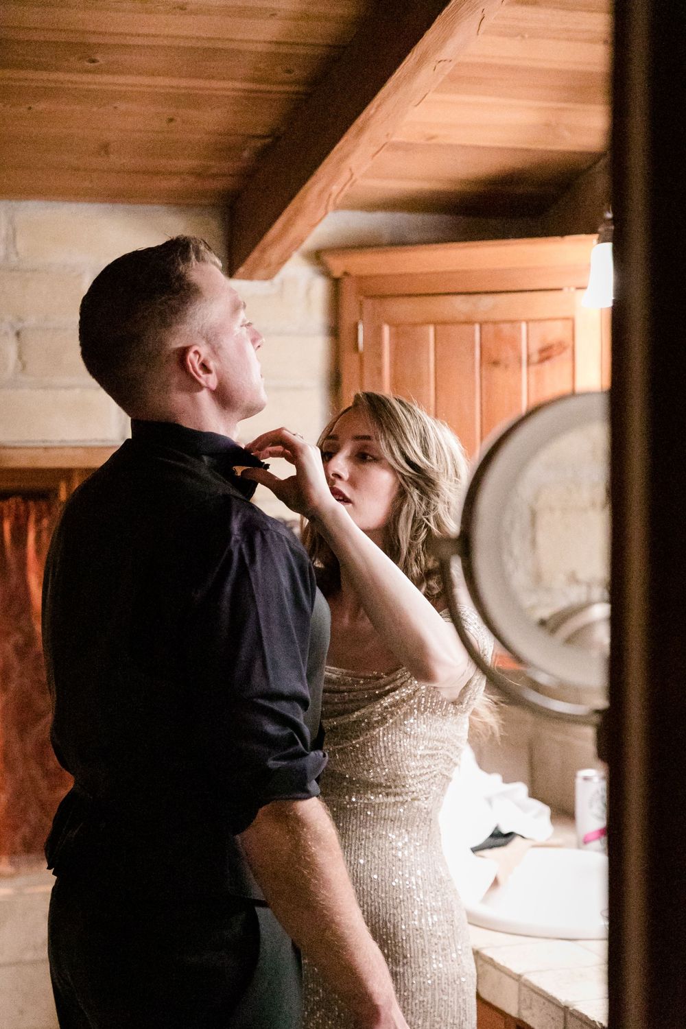 A woman in a gold sequin dress adjusts a man's bow tie in a rustic cabin bathroom.