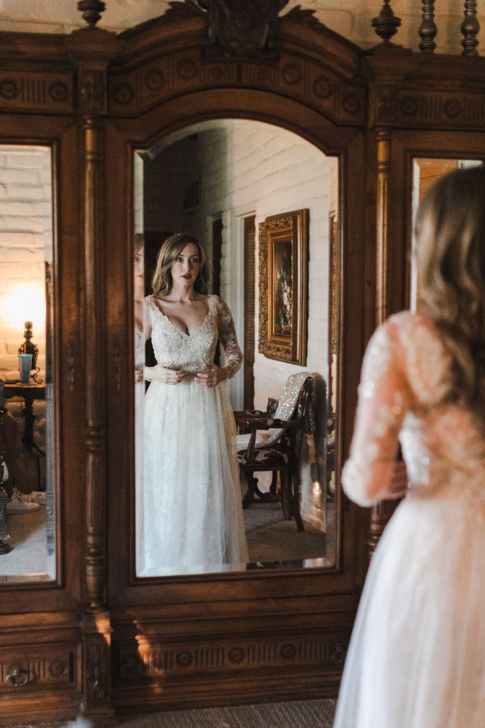 Bride in long-sleeve lace wedding dress admiring her reflection in an ornate wooden mirror.