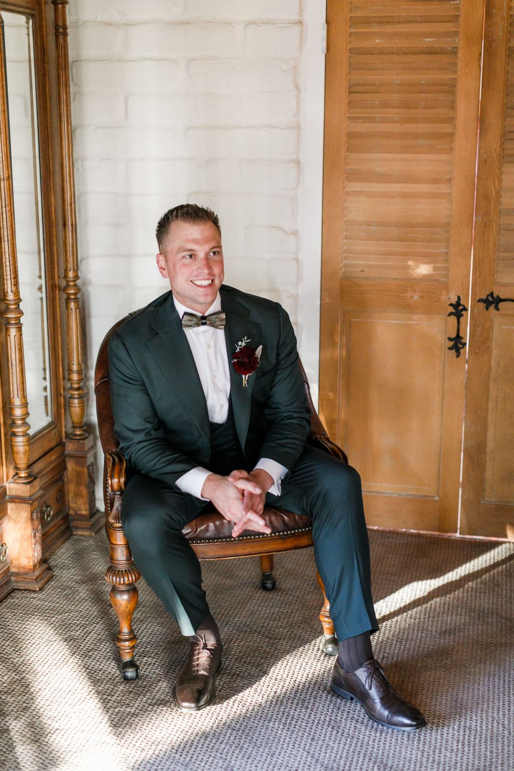 Smiling groom in dark green tuxedo with bow tie and boutonniere sits in vintage chair by wooden doors.