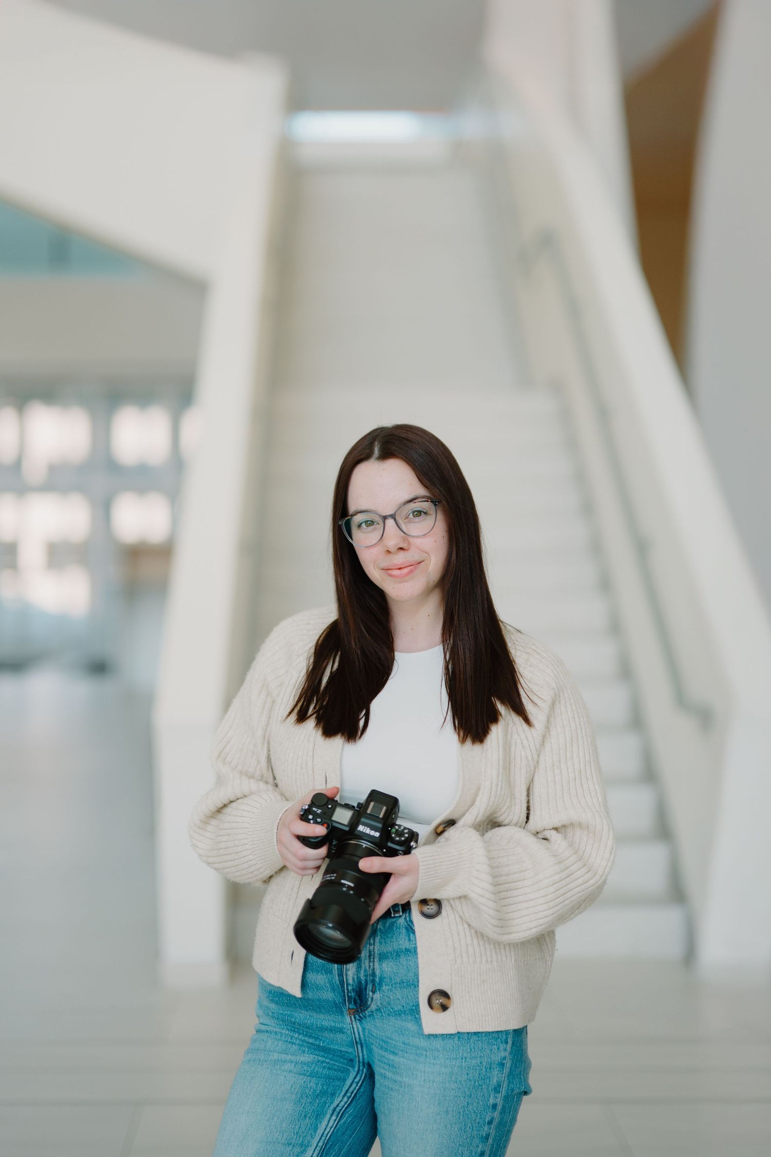 Woman in glasses and cream sweater poses with camera in modern architectural setting with white columns.