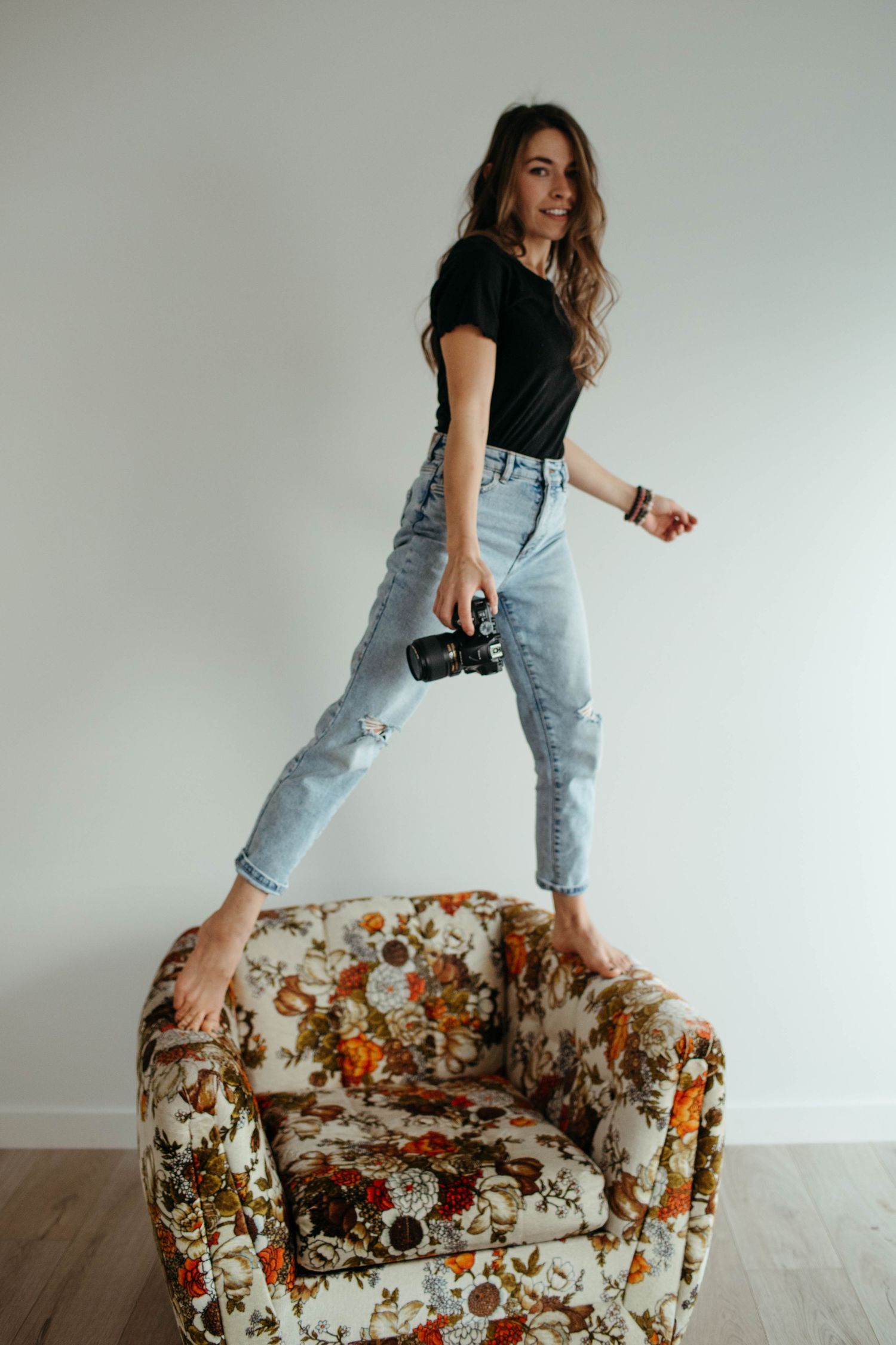 Young woman in casual wear standing playfully on floral armchair against white wall.