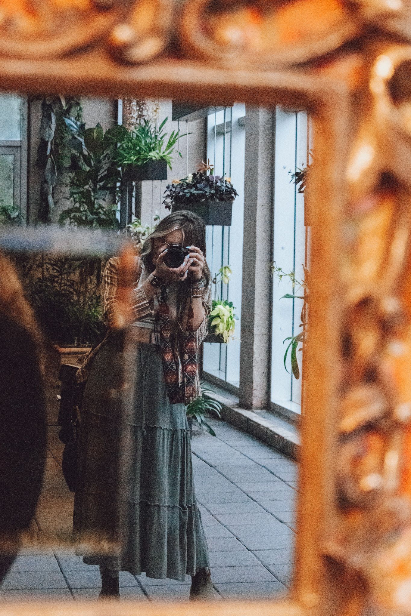 Artistic reflection in mirror shows someone taking a photo among indoor plants by a window.