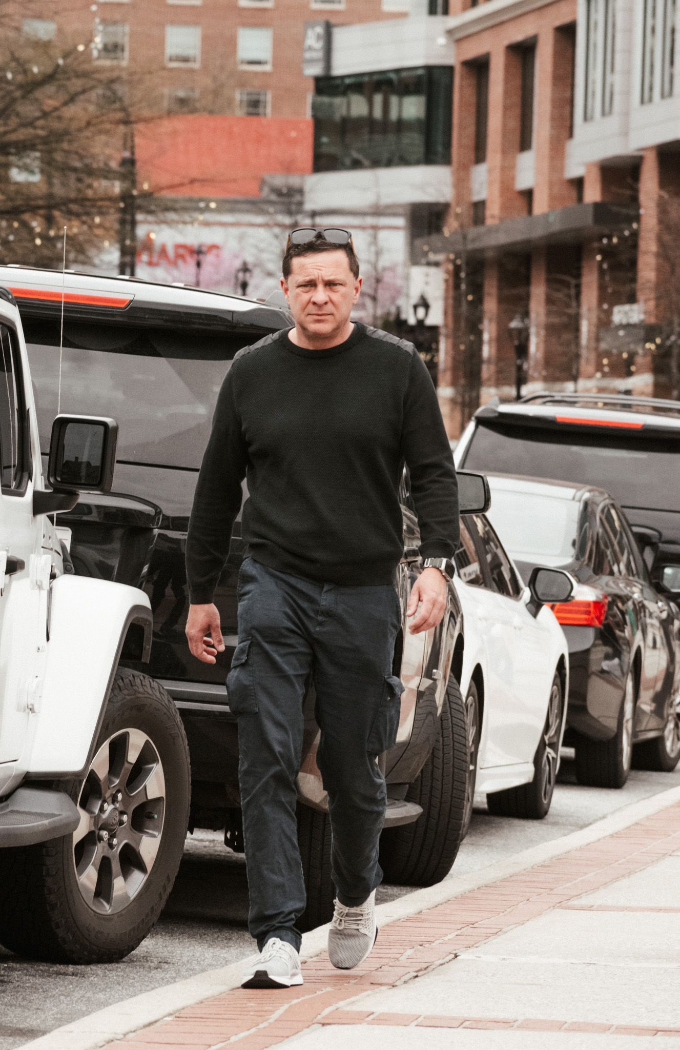 Man in black sweater and dark pants walking on urban sidewalk past parked cars.