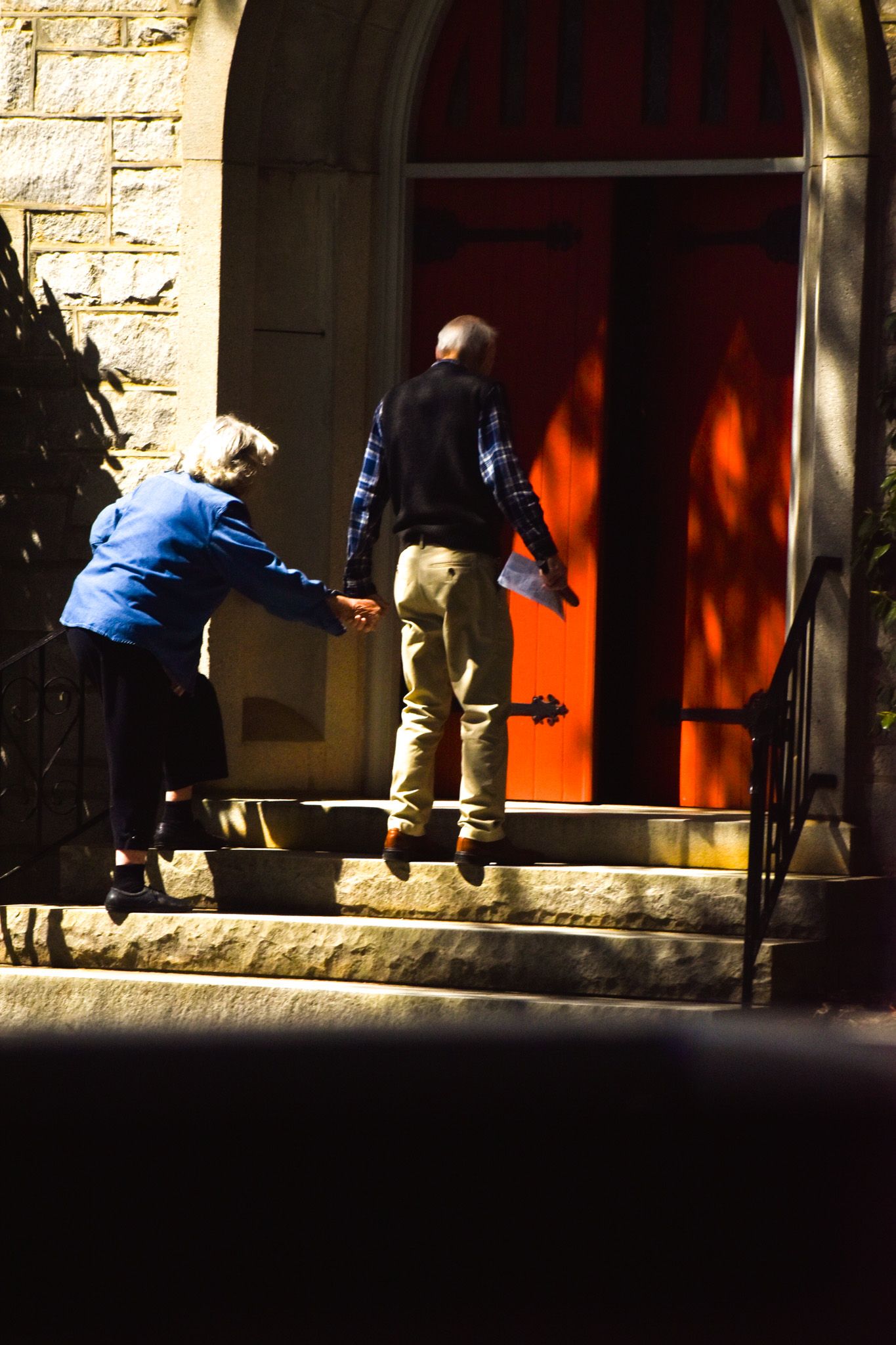 Two people in casual attire stand at a doorway with a bright red door and stone facade.
