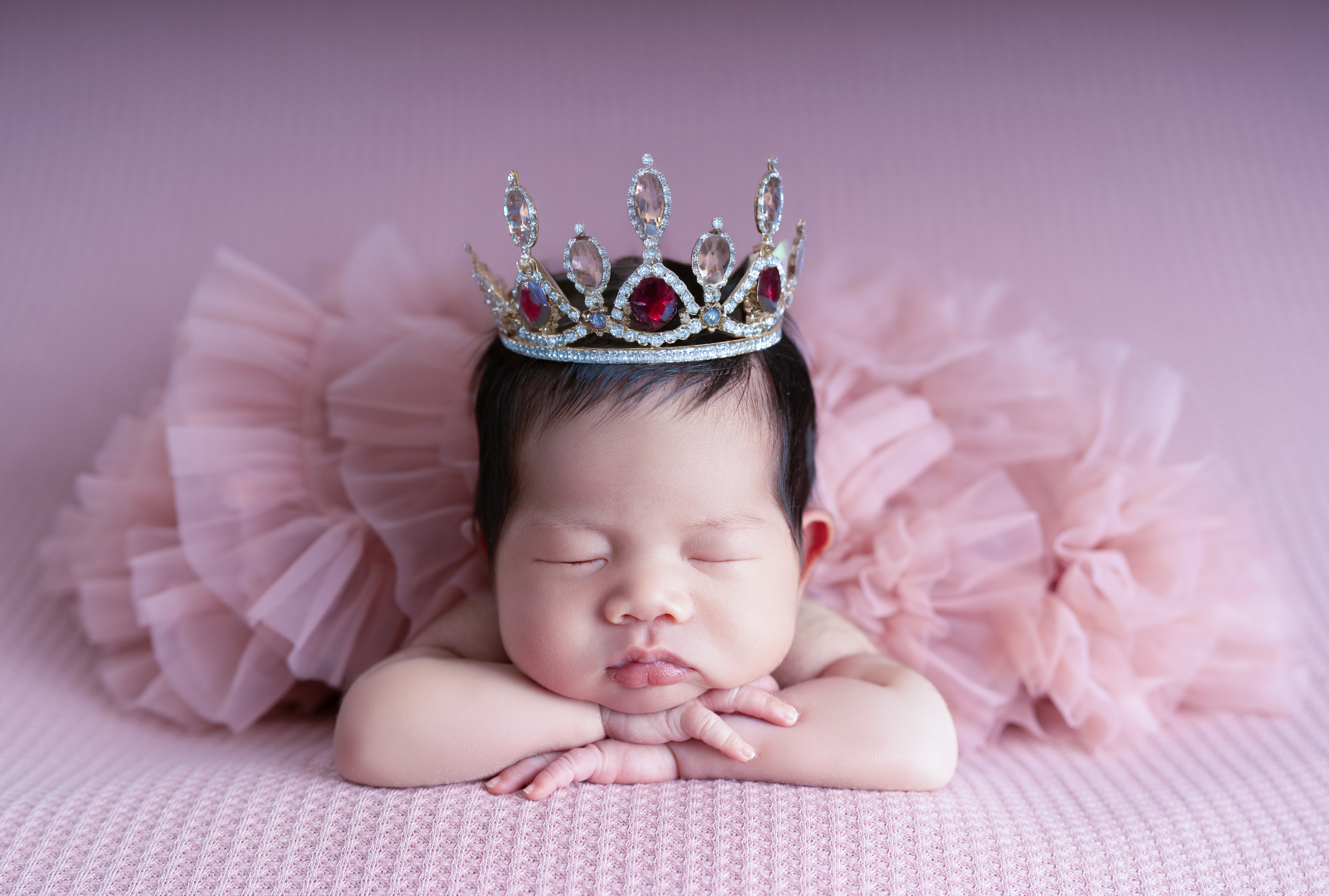 A sleeping newborn baby wearing a pink tutu and a jeweled crown poses with hands under chin on a pink background.