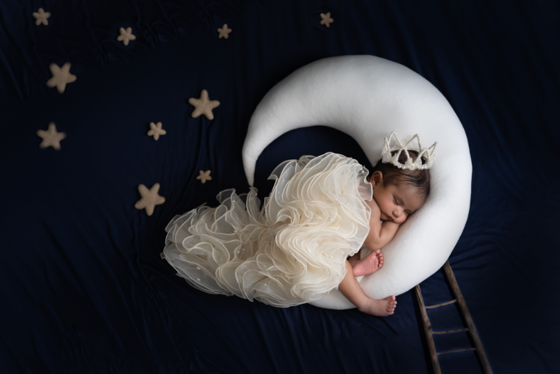 Newborn baby girl in white ruffled dress sleeping on crescent moon prop with crown, surrounded by stars on navy background. Embracing Life’s