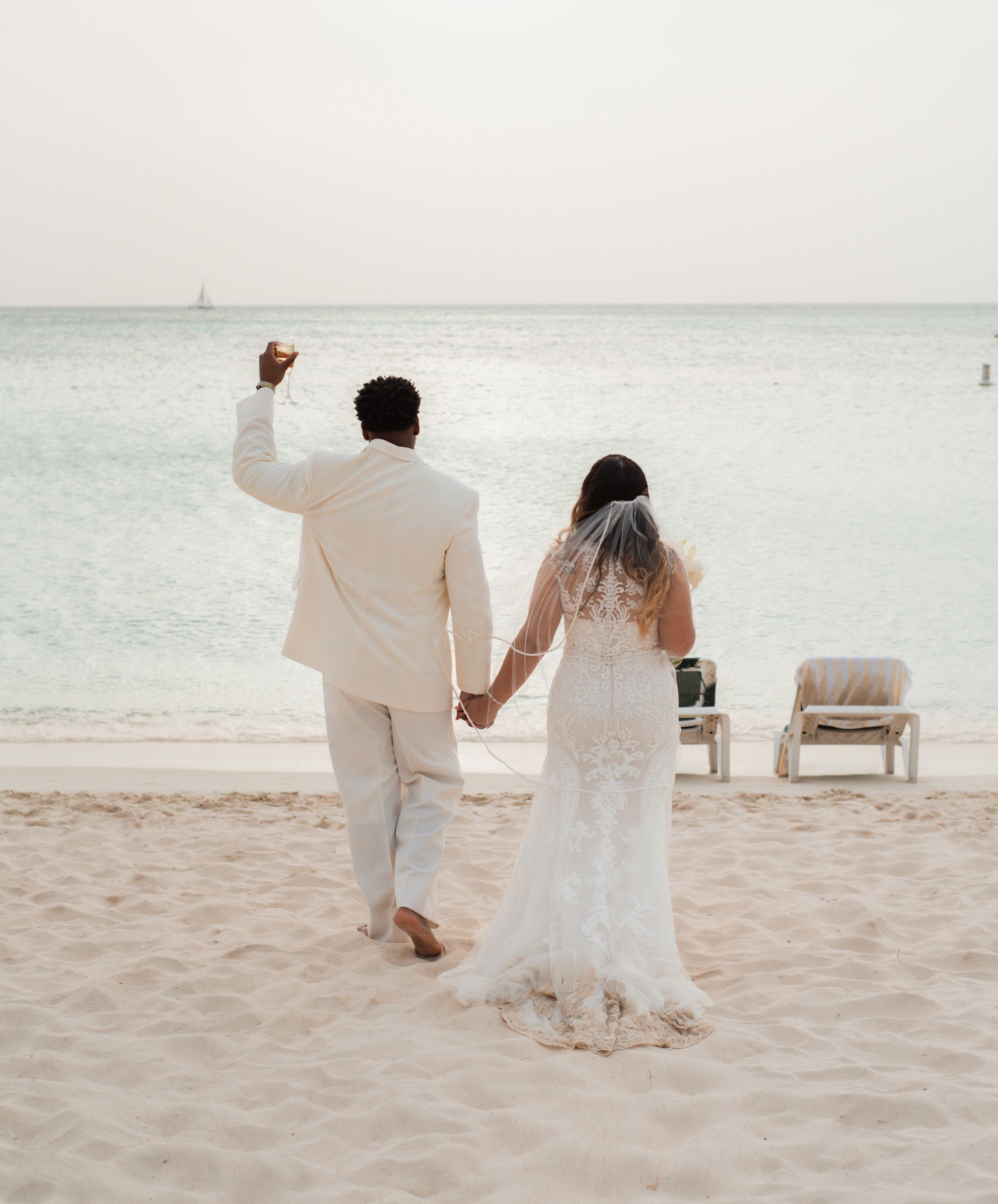 A couple in white attire celebrates on a beach with boats visible in the distance.