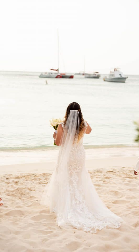 A bride in a lace wedding dress with flowing veil stands on a beach near sailboats.