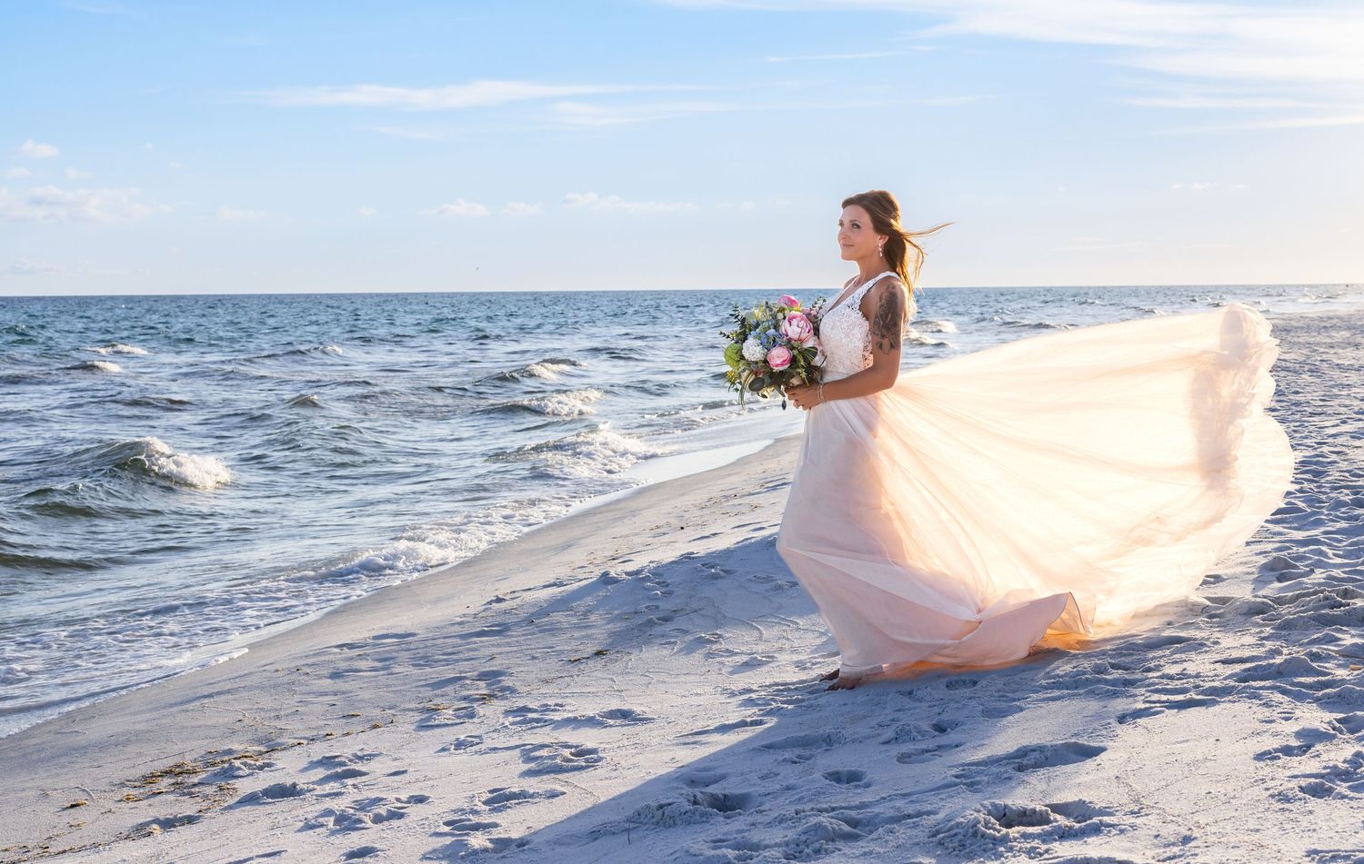 Wedding dress flows in beach breeze as bride holds bouquet by ocean waves.
