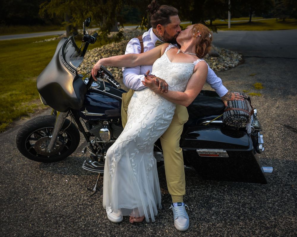 Newlywed couple kissing in wedding attire beside a black Harley-Davidson motorcycle outdoors.