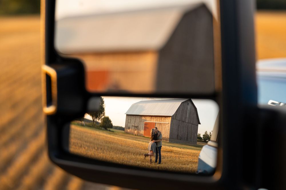 A couple embraces near a barn, reflected in a truck side mirror during golden hour on a farm.