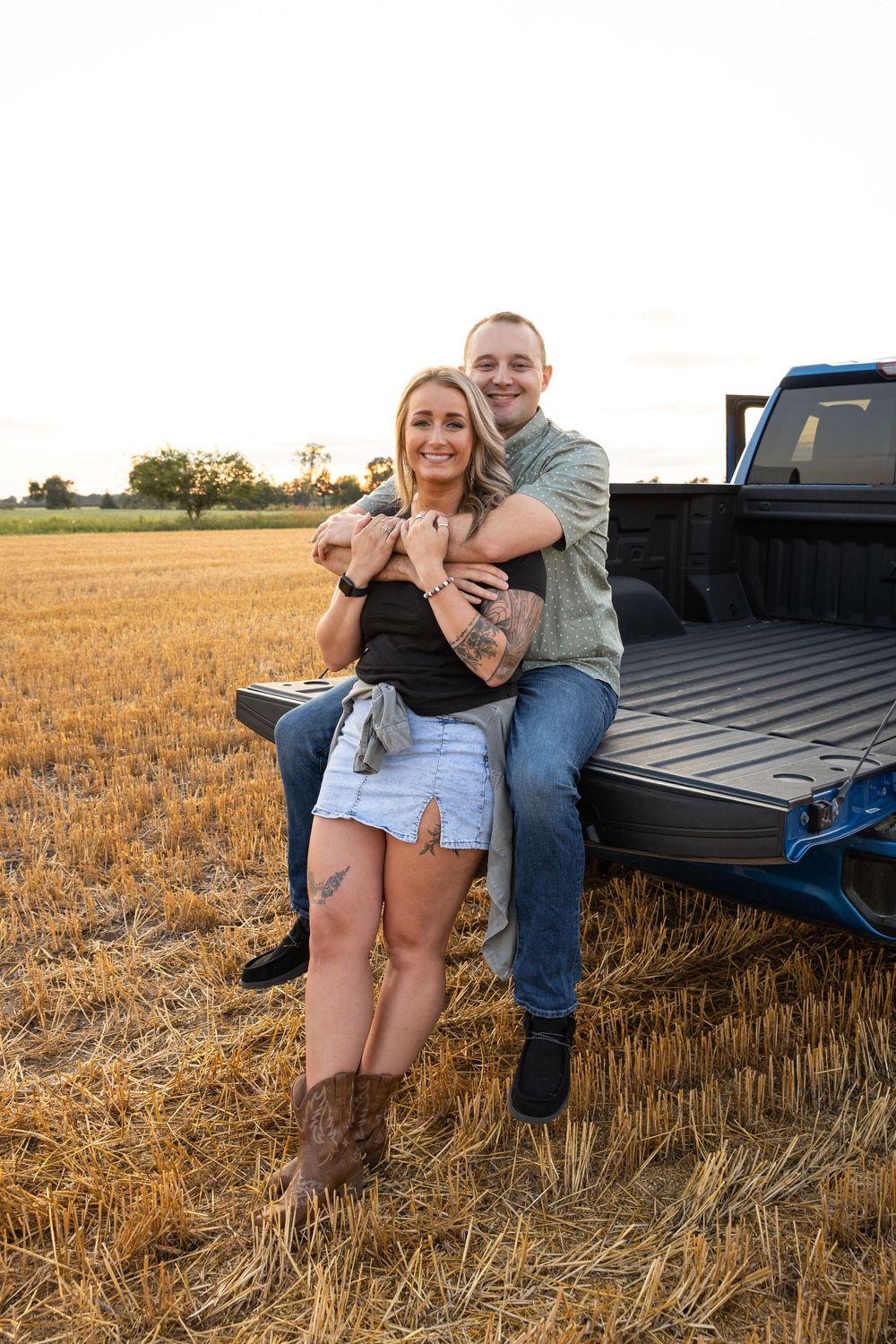 Couple embracing on a pickup truck tailgate in a golden hay field at sunset, woman in denim shorts and cowboy boots.