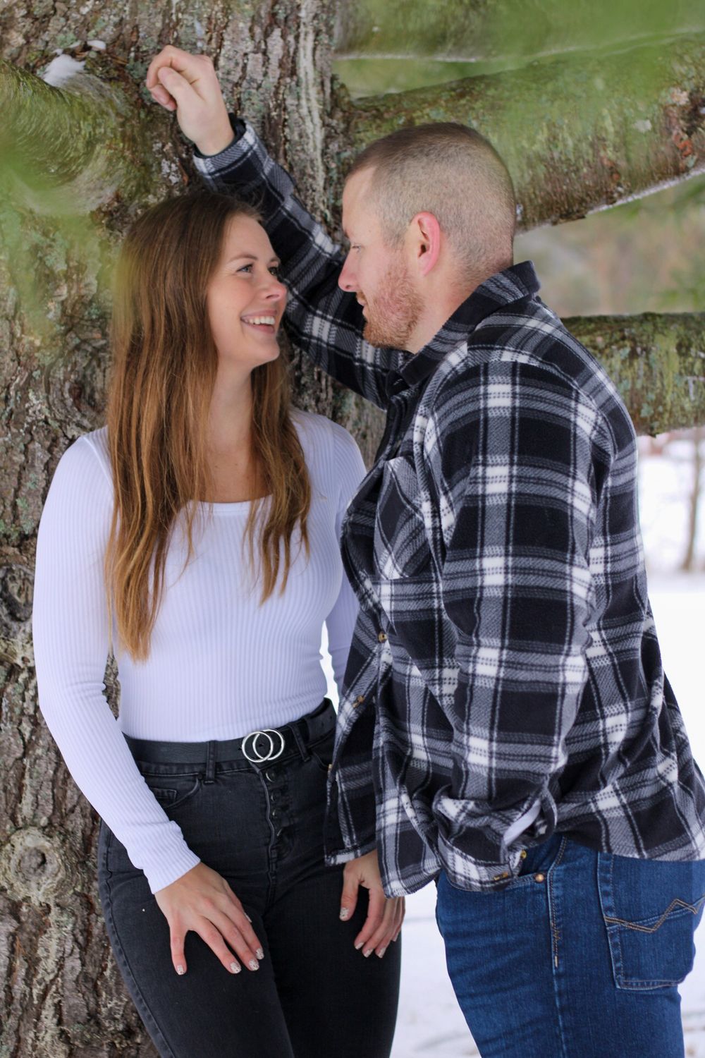 Couple smiling at each other leaning against a tree in a snowy winter setting, woman in white top and man in plaid shirt.
