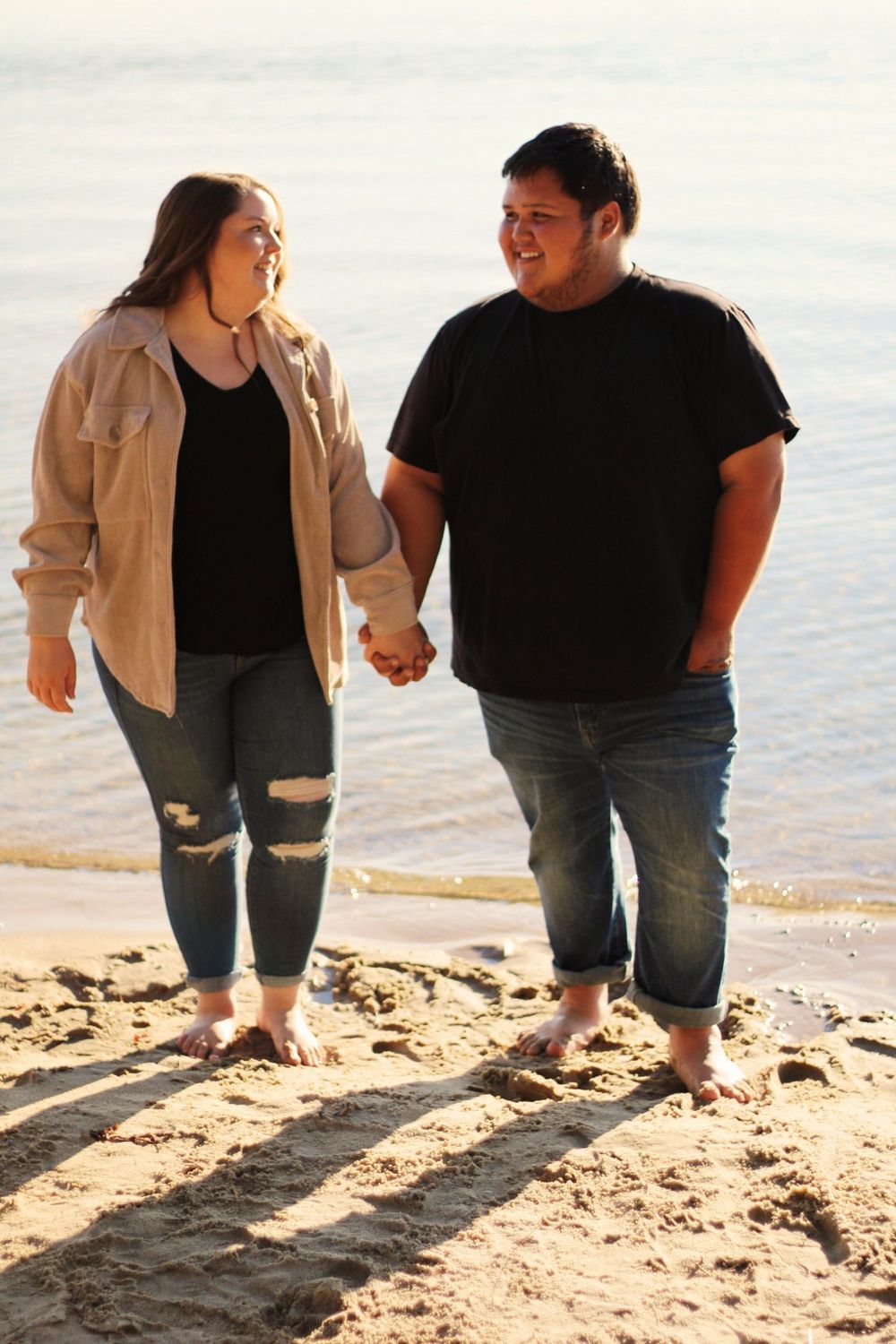 Couple holding hands and walking barefoot on a sandy beach near the water's edge at sunset.