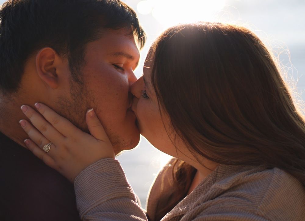 Couple sharing a romantic kiss outdoors with warm golden backlight, woman wearing an engagement ring.