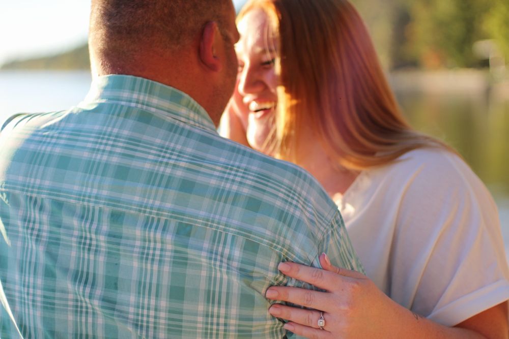 Couple embracing outdoors near a lake, woman smiling and wearing an engagement ring, in warm golden light.