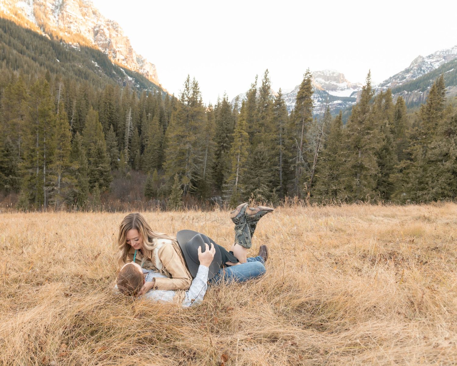 Couple lying playfully in tall golden grass with snow-capped mountains and evergreen forest in the background.