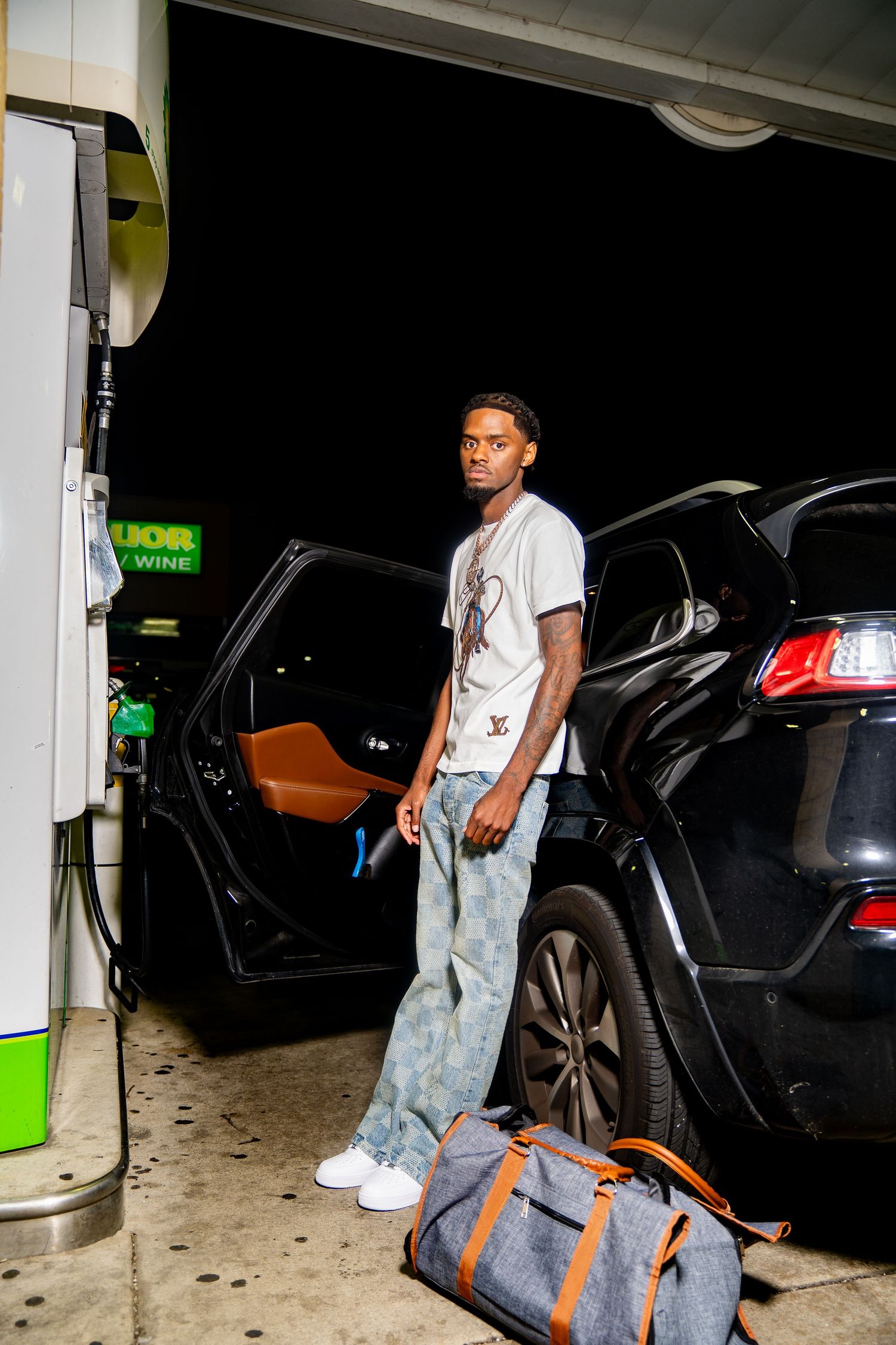 Person standing at gas station at night next to black SUV with travel bag.