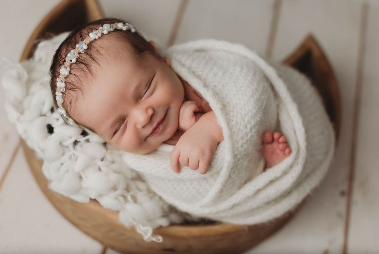Sleeping newborn baby wrapped in soft beige swaddle with floral headband on light background.