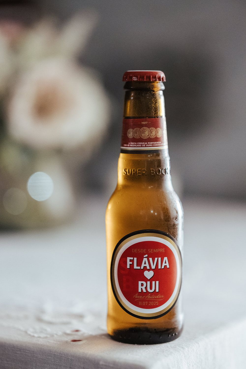 Custom Super Bock beer bottle labeled Flávia e Rui at a Portuguese wedding reception with floral decor in background.