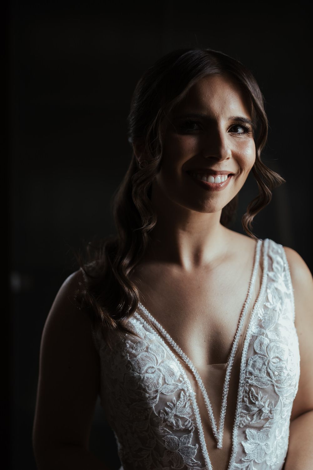 Radiant bride in deep V-neck lace wedding dress smiling softly in dramatic dark studio portrait lighting.