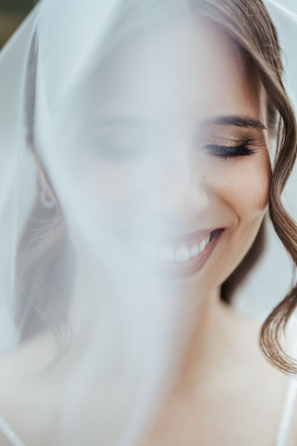 Beautiful bride smiling joyfully beneath a sheer white wedding veil in a dreamy soft-focus bridal portrait.