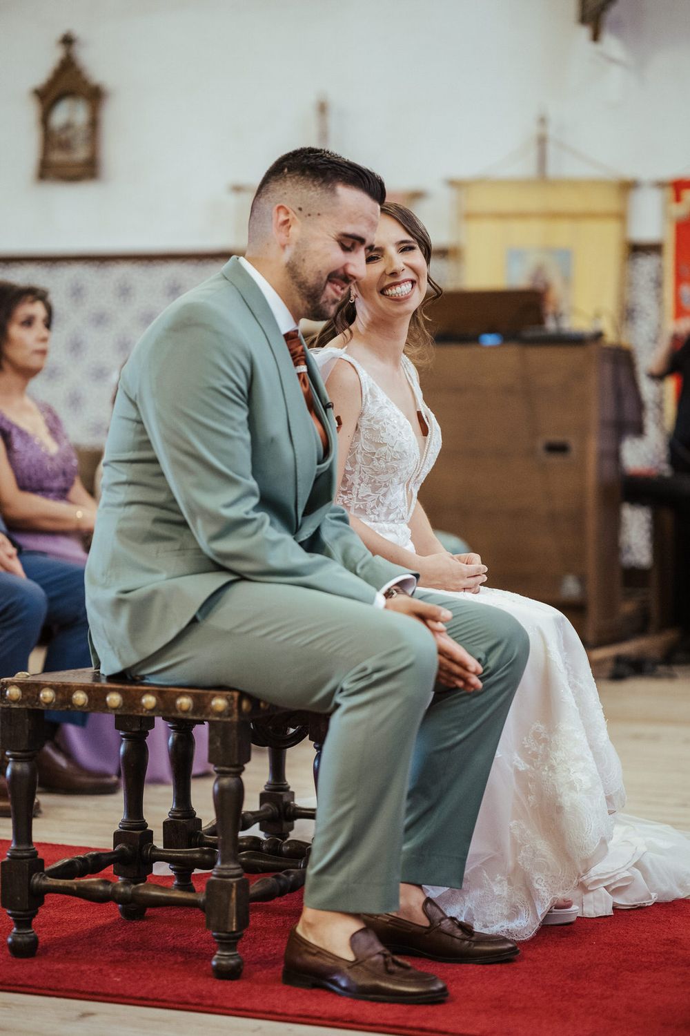 A joyful bride in a lace gown and groom in a sage suit laugh together while seated during their church wedding ceremony.