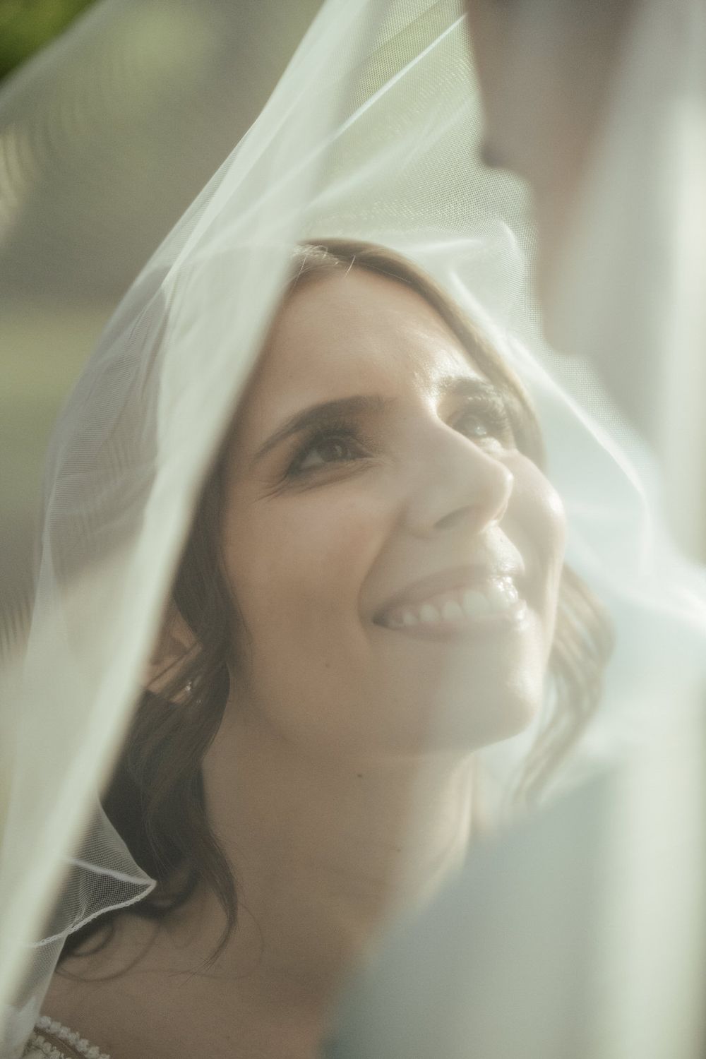 Radiant bride smiling under sheer white veil in soft natural light on her wedding day.