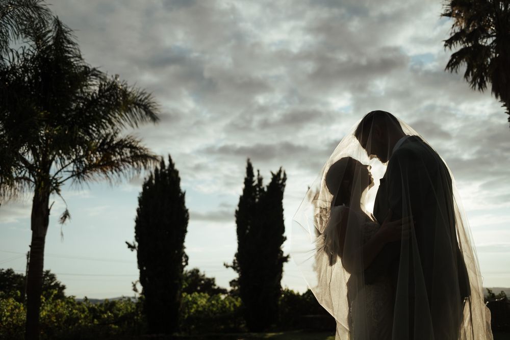 Romantic silhouette of bride and groom sharing an intimate moment under dramatic cloudy sky at sunset.