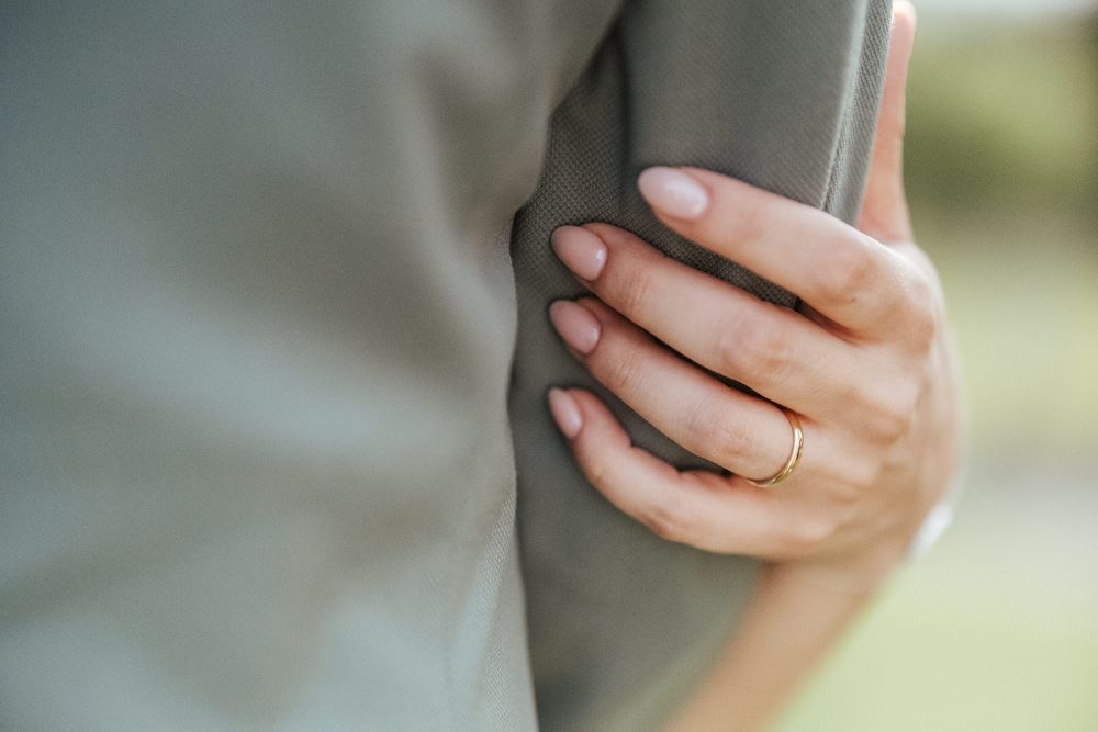 Close-up of a bride's hand with a gold wedding ring gently holding the groom's arm in a grey suit.