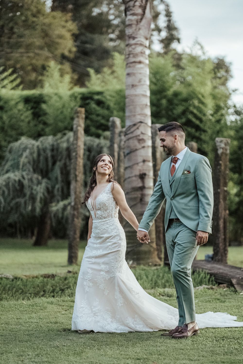 Happy bride and groom holding hands in lush garden, groom wearing sage green suit with coral tie.