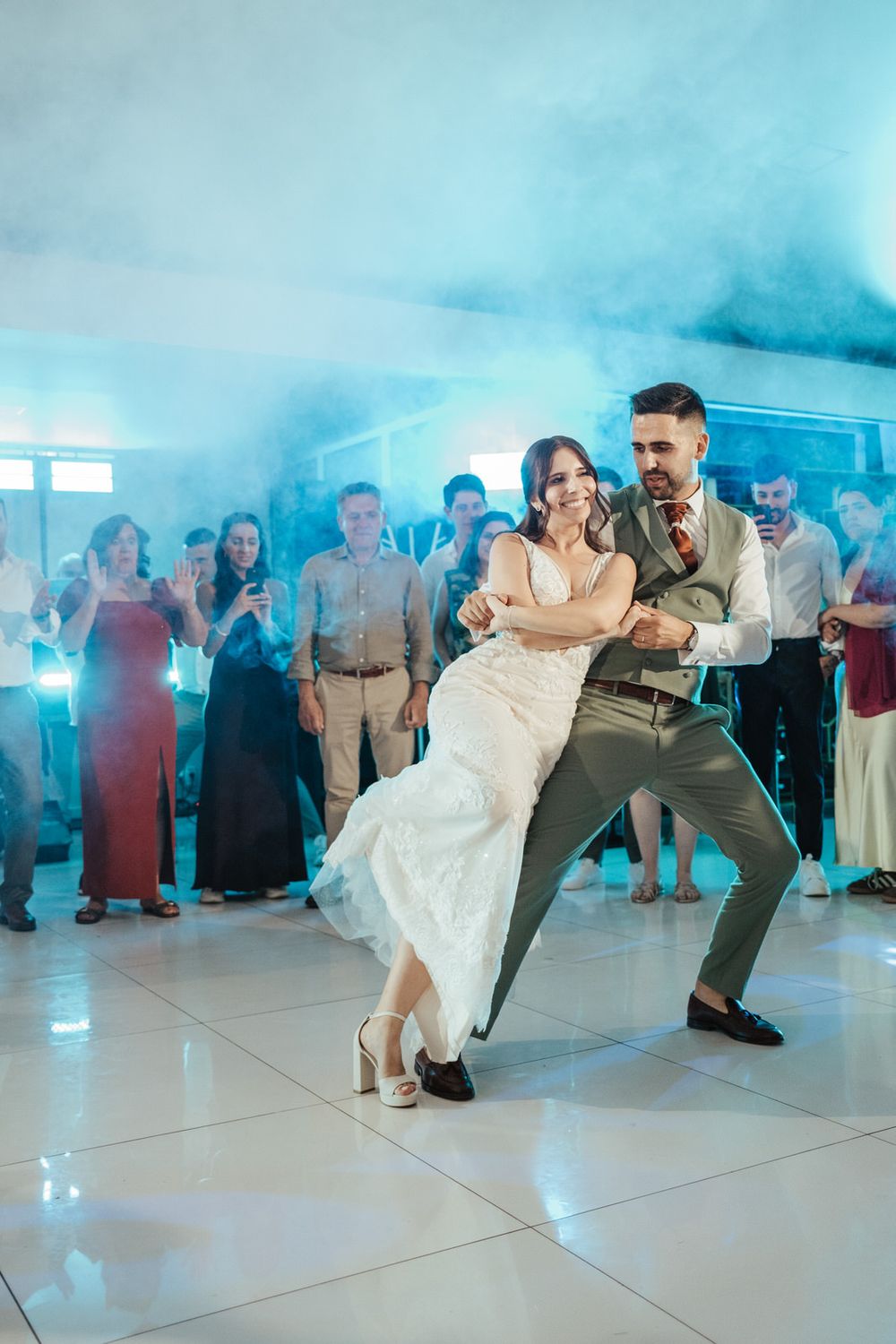 Bride and groom share their first dance on a white dance floor surrounded by guests and blue atmospheric lighting.