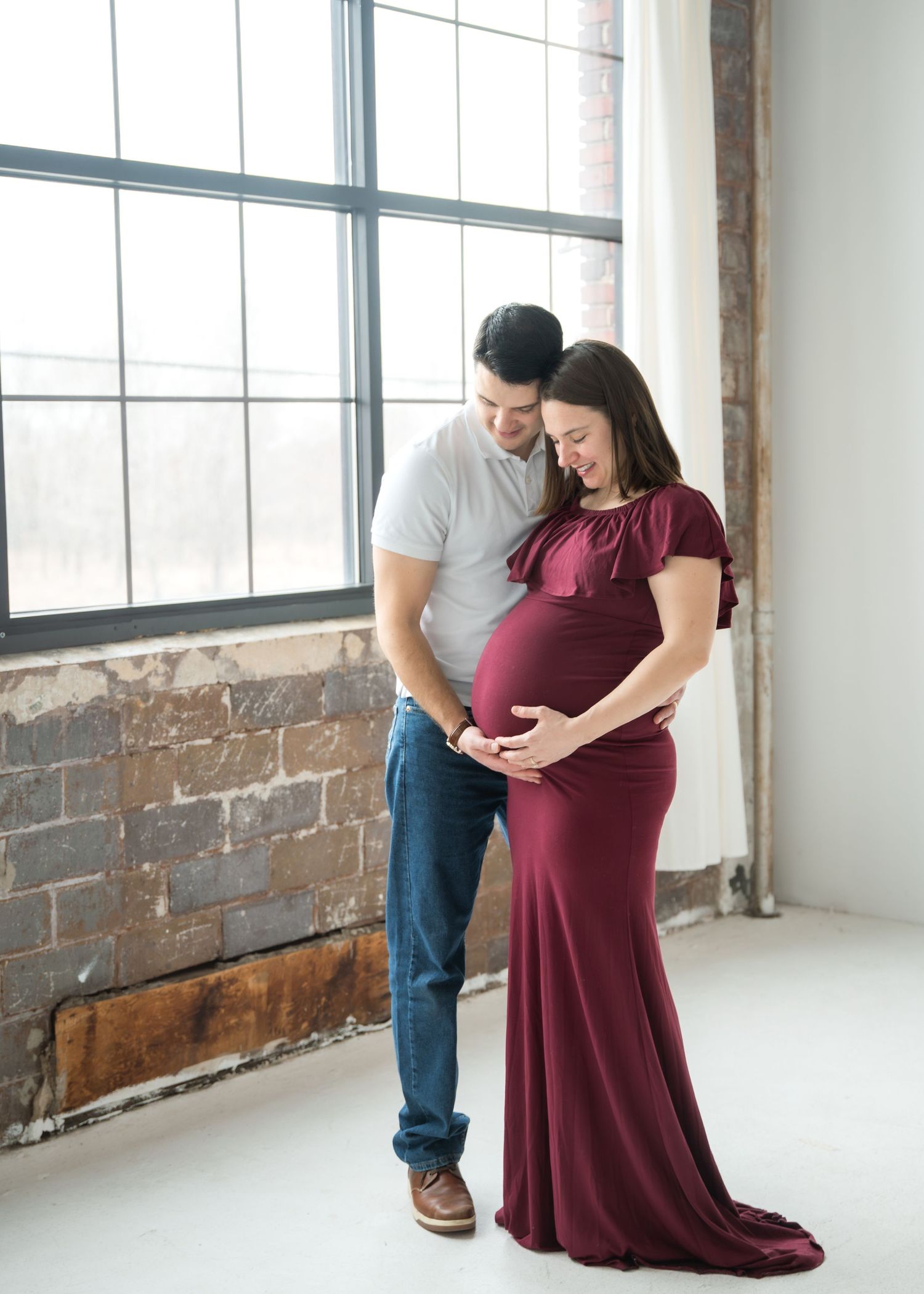 Couple sharing a tender moment during maternity photoshoot, woman in burgundy gown cradling baby bump by large window.