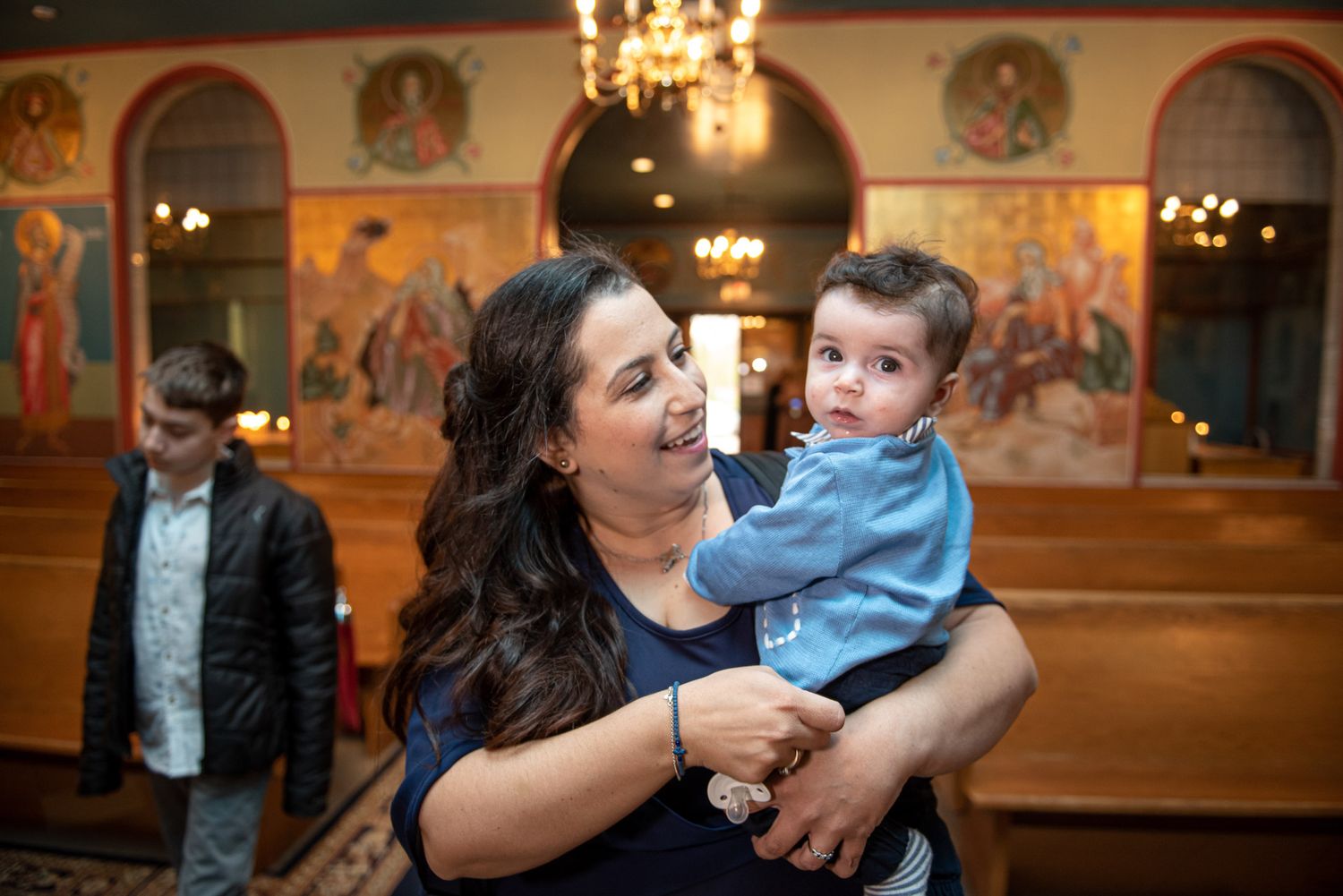 Mother holding baby boy in blue outfit inside decorated Orthodox church with murals.
